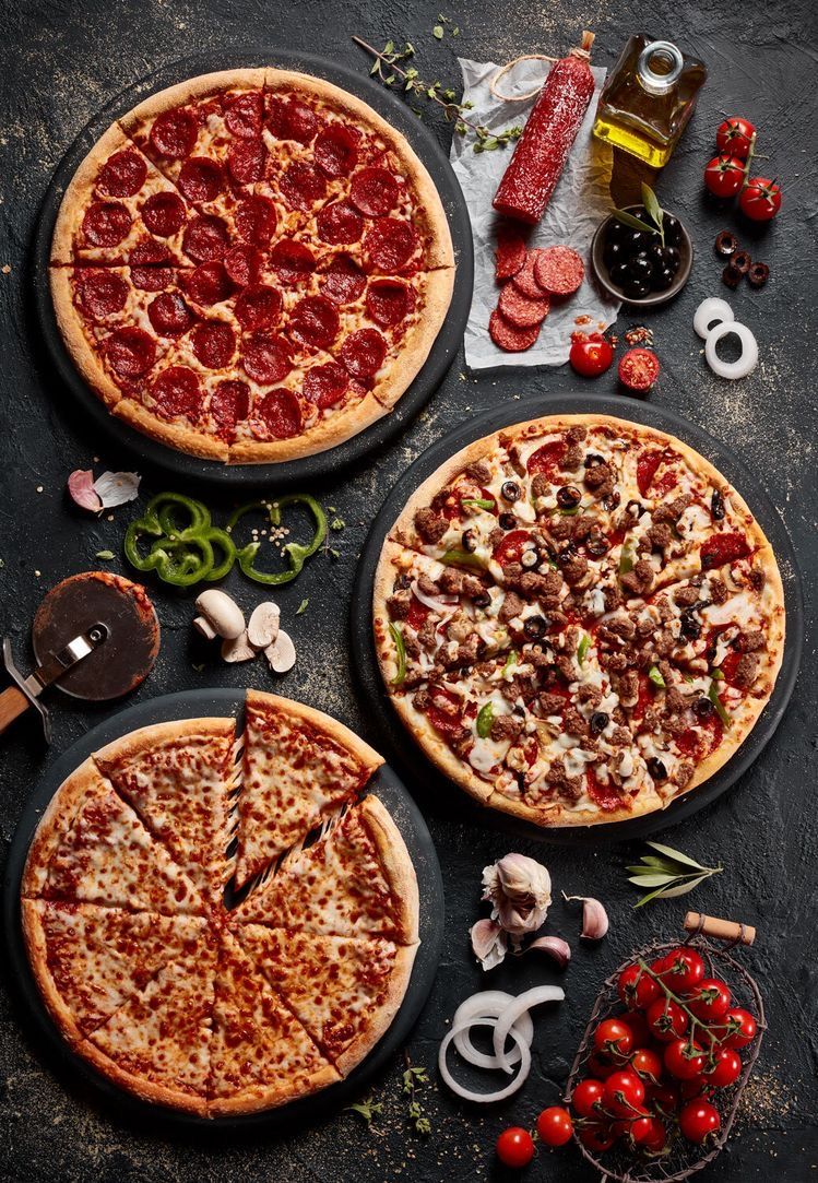 Three richly topped pizzas arranged on a dark tabletop with ingredients around them.